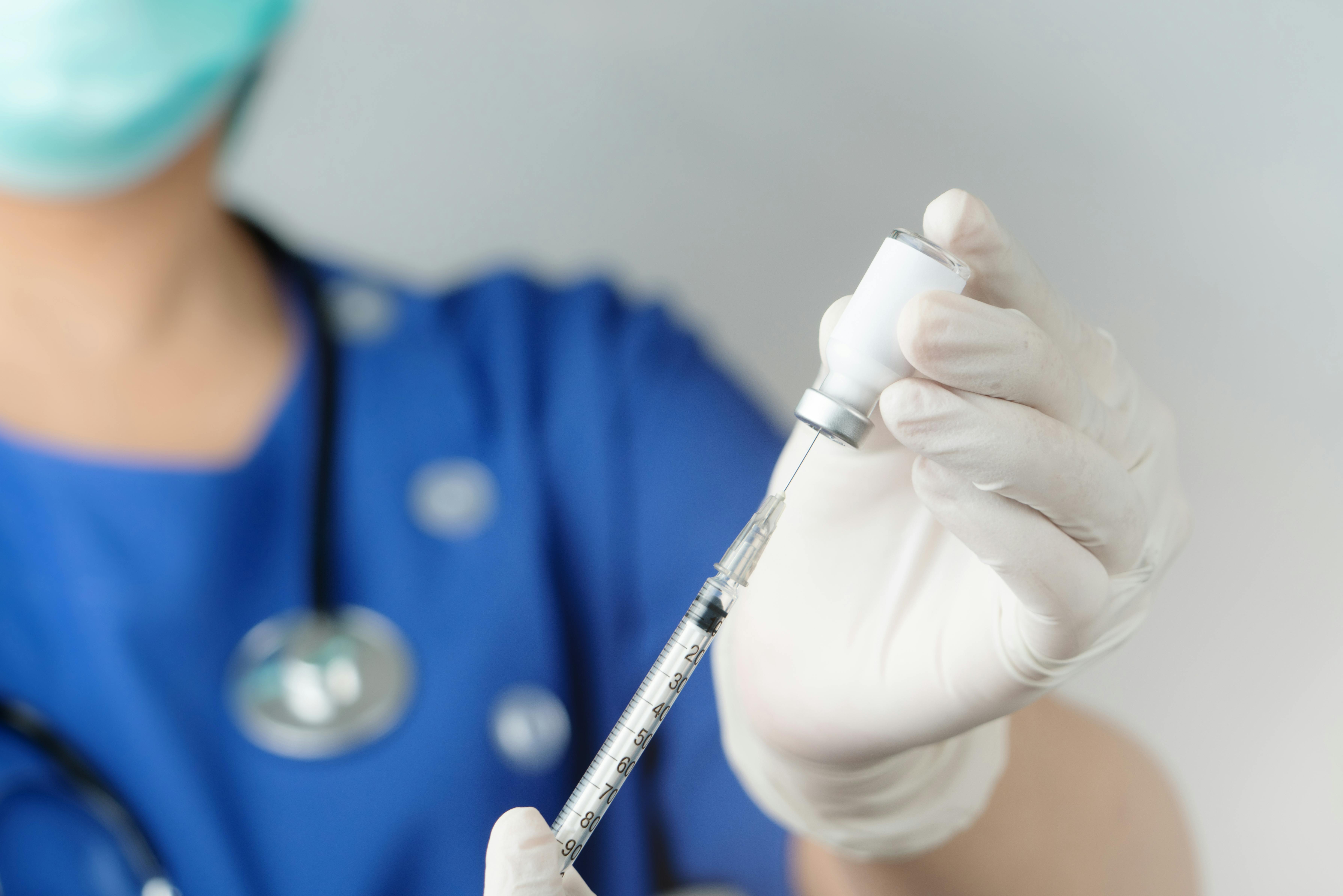 Person in hospital scrubs and mask filling a vaccine needle from a bottle.